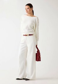 Pull blanc en tricot avec détails latéraux, associé à un pantalon blanc à jambes larges. Accessoirisé avec un sac à main rouge et une ceinture assortie.