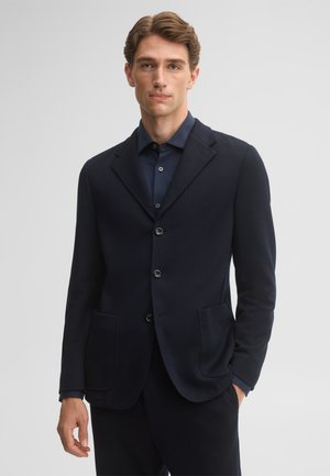 Jonge man draagt een donkerblauw colbert over een bijpassend donkerblauw overhemd met knopen, staand voor een effen lichtgrijze achtergrond.