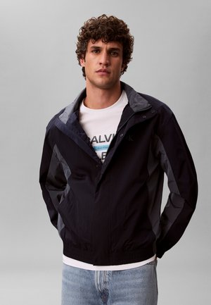 Jeune homme aux cheveux bouclés portant une veste noire et grise par-dessus un t-shirt blanc Calvin Klein et un jean bleu clair, debout devant un fond gris.