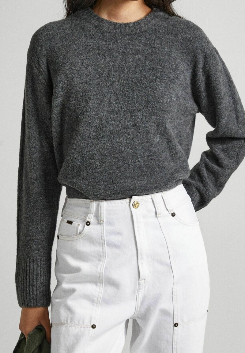 Femme portant un pull à manches longues gris foncé rentré dans un pantalon blanc taille haute, tenant un tissu vert dans sa main gauche.