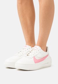 Nohy v bílých teniskách Nike Air Force 1 s růžovým logem Swoosh, stojící na bílém pozadí.