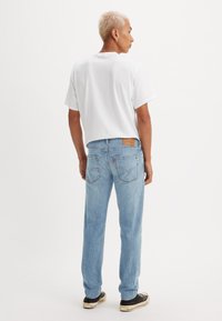 Bărbat cu păr scurt blond, purtând un tricou alb, blugi deschiși la culoare și adidași negri, stând cu spatele pe un fundal alb.