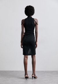 Vestido negro ajustado con cuello alto, detalle fruncido y tirantes ajustables. Terminación con un dobladillo a la altura de la rodilla y un panel semitransparente en la parte posterior.