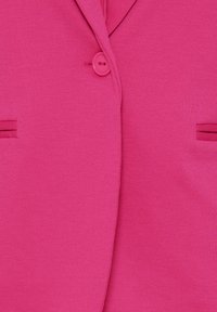 Gros plan d'un blazer rose vif avec un bouton assorti fermé et deux poches horizontales fendue de chaque côté.