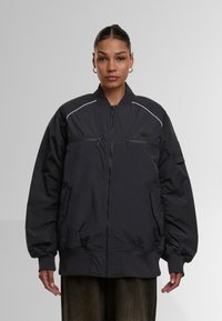Moteris vilki didelį juodą zip-up bomber tipo švarką su raukšlėtomis rankovėmis ir hemu, stovi prieš paprastą šviesiai pilką foną.