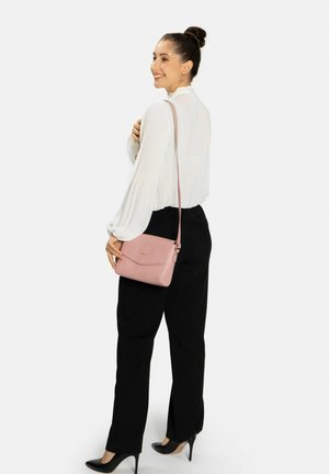 Bolso cruzado de cuero rosa con acabado texturizado, cuenta con una tapa frontal y una correa larga ajustable. Llevado por un modelo con pantalones negros.