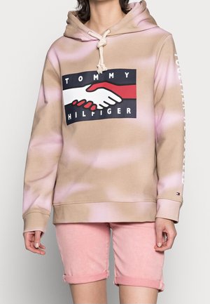 Personne portant un sweat à capuche beige et rose tie-dye avec un logo poignée de main et le texte "Tommy Hilfiger", associé à un short rose retroussé.