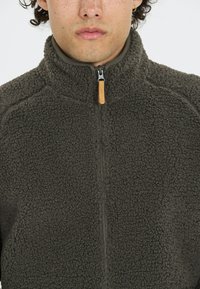 Fleece zipjacka i mörkgrått med hög krage, texturerad yta och en dragkedja med trädetalj. Har en tight passform.