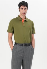 Grønn polo med oransje krage og logo, kombinert med grå stripete bukser og svart belte. Korte ermer og klassisk passform.