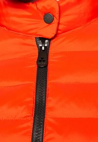 Veste matelassée orange vif dotée d'une fermeture éclair noire et d'un bouton-pression au niveau du col, avec des coutures horizontales pour l'isolation.