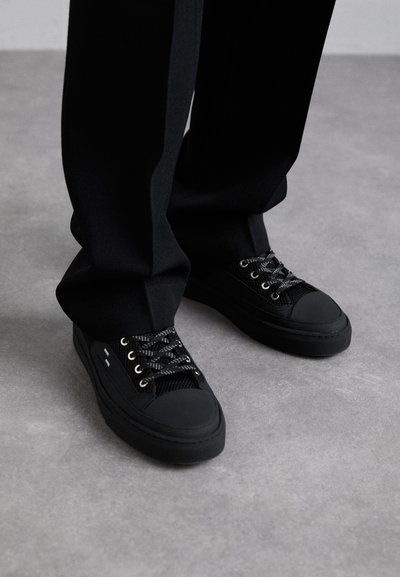 Schwarze Sneakers mit einem Obermaterial aus Netzgewebe, Gummi-Zehen, Metallösen und kontrastierenden Schnürsenkeln. Kombiniert mit schwarzen Hosen auf einer grauen Oberfläche.