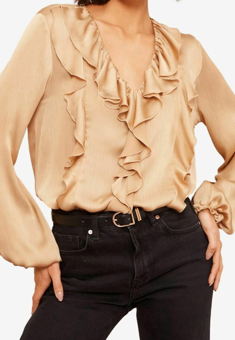 Blouse beige avec un col et des manches volants, confectionnée dans un tissu lisse. Associée à un jean noir et une ceinture noire avec une boucle en or.