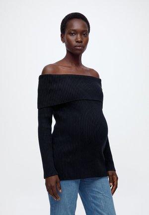 Femme enceinte portant un pull noir côtelé à épaules dénudées et un jean bleu, debout devant un fond blanc uni.