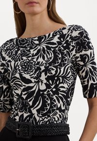 Woman wearing black and white floral patterned top with elbow-length sleeves and a black woven belt, shown from shoulders to waist.