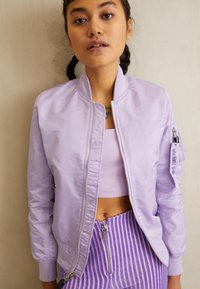 Levandulová bomber bunda so zipsom v prednej časti, rebrovanými manžetami a bočnou vreckom. Vnútorná podšívka je hladká; nosí sa cez zodpovedajúci crop top.