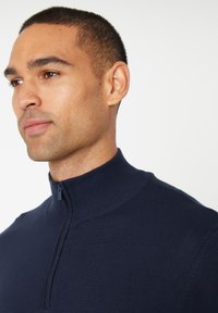 Navyblauwe ritstrui met hoge kraag, gemaakt van een gladde, gebreide stof. Bevat een korte rits bij de nek en ribgebreide boorden.