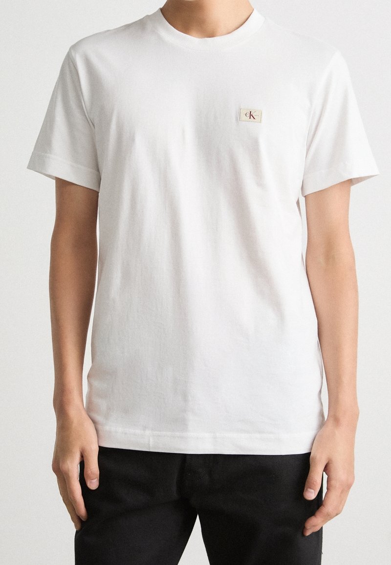 T-shirt bianco a maniche corte con collo a giro, con piccolo patch del logo Calvin Klein sul lato sinistro del petto, indossato da una persona con pantaloni neri.