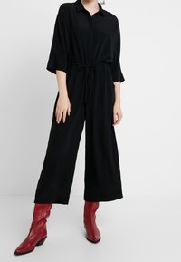 Černý overal s knoflíkovým zapínáním vpředu, širokými rukávy a stáhnutým pasem. V kombinaci s červenými koženými kovbojskými botami.