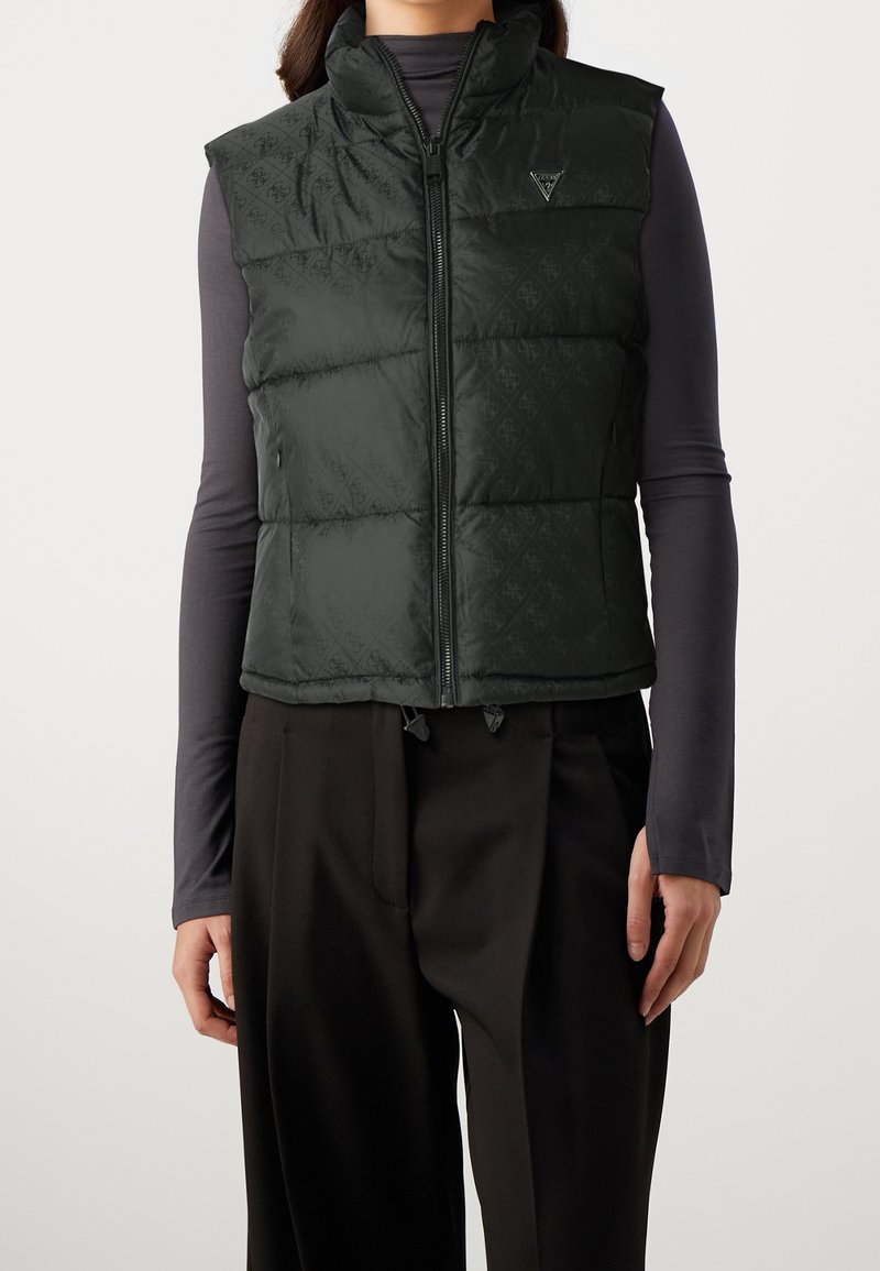 Donna che indossa un gilet imbottito senza maniche nero trapuntato con una zip frontale sopra una maglia a maniche lunghe grigio scuro e pantaloni neri.