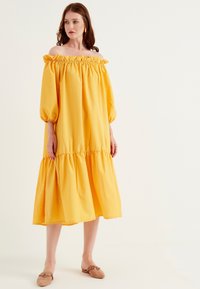 Gul off-shoulder klänning med volangkrage, puffärmar och lager-på-lager kjol. Slätt tyg och knälång design.