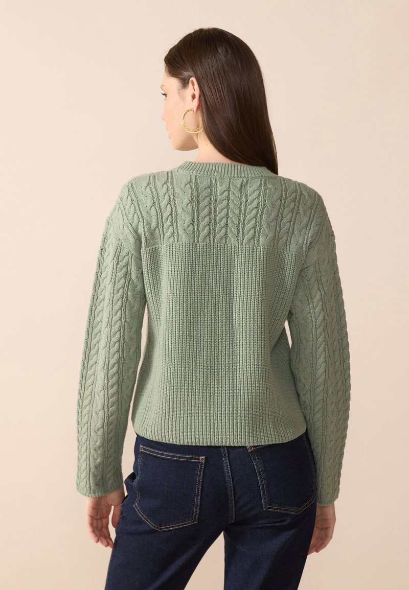 Maglione lavorato a maglia verde con un design a trecce testurizzato, orlo a coste e maniche lunghe, abbinato a pantaloni in denim scuro.