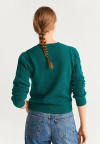 Pull tricot bleu sarcelle avec manches bouffantes texturées, poignets et ourlet côtelés. Associé à un jean bleu taille haute, présentant une coupe décontractée.