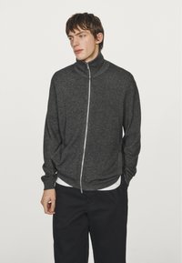 Grå zip-up jacka med hög krage, texturerad tyg, långa ärmar och sidofickor, bärs över en vit skjorta och matchas med mörka byxor.