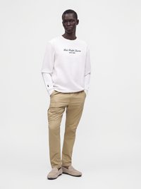 Άνδρας που φοράει λευκό φούτερ Polo Ralph Lauren, μπεζ παντελόνι cargo και μπεζ μοκασίνια, στέκεται με τα χέρια στις τσέπες σε απλό φόντο.