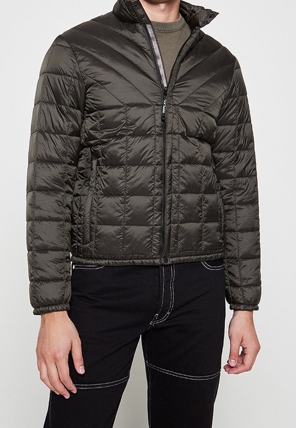 Homme portant une doudoune matelassée vert foncé par-dessus une chemise verte et un pantalon noir avec des coutures blanches, debout sur un fond uni.