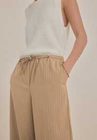 Beige gestreifte Hose mit elastischem Bund und Kordelzug, kombiniert mit einem weißen ärmellosen Strickoberteil, das eine weiche Textur zeigt.