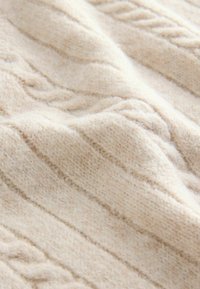 Strickstoff in hellem Beige mit strukturierten Zöpfen und Rillen, der eine weiche, flauschige Textur mit feinen Tiefen- und Musterdetails betont.