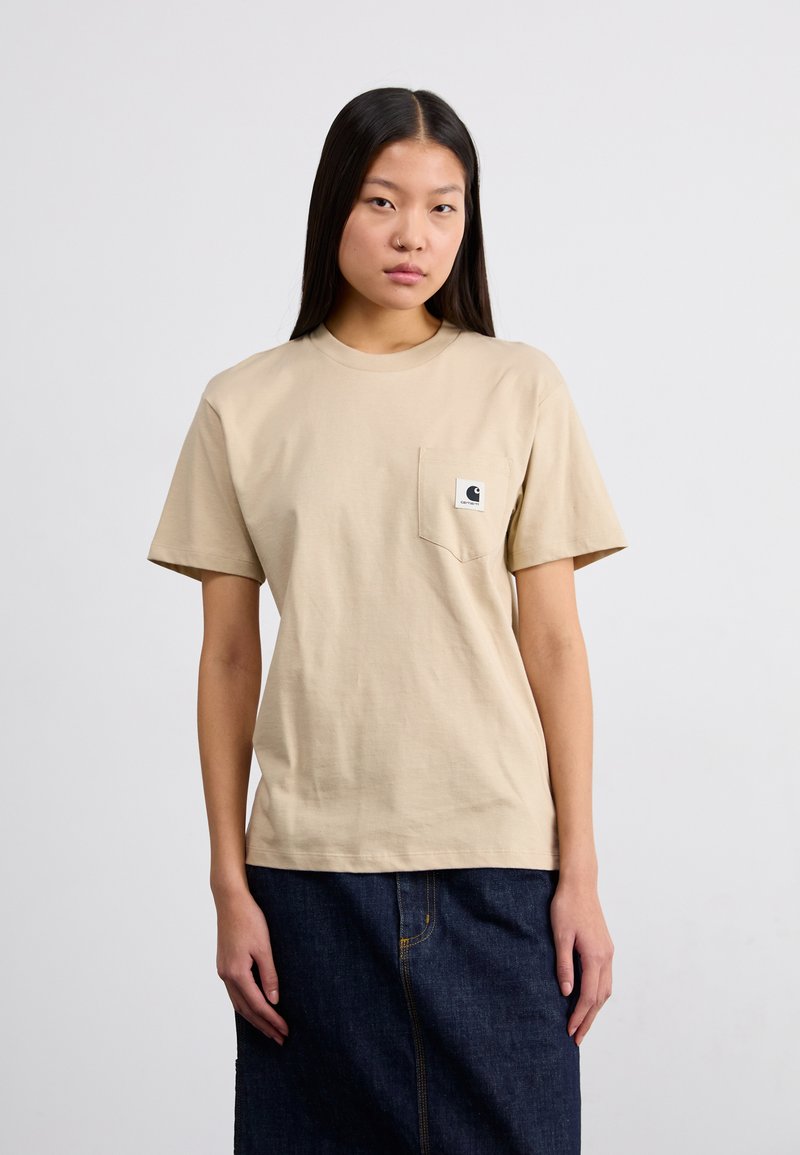 T-shirt en coton beige avec col rond, manches courtes, et poche sur la poitrine gauche arborant un écusson. Associé à une jupe en denim foncé.