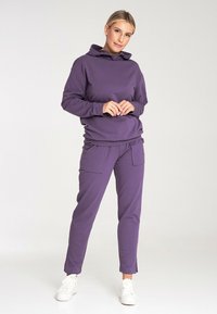 Fioletowy hoodie i dopasowany komplet dresowy wykonane z miękkiego materiału; posiadają zakładany kaptur, boczne kieszenie oraz zwężane spodnie z rozcięciami na kostkach.