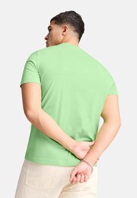 Maglietta verde a girocollo in cotone, maniche corte, texture liscia, vestibilità comoda, indossata con le mani incrociate dietro la schiena.