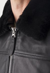 Svart läderjacka med en stor, mjuk svart fårskinnskrage. Har en silverblixtlås, texturerat läder och horisontell sömdekor.