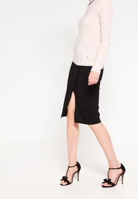 Svart knälång kjol med en frontslits, i kombination med en ljusrosa långärmad tröja. Svarta högklackade sandaler med rosettdetaljer.