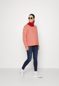 Pulôver de malha oversize listrado em vermelho e branco com um gola alta. Combinado com jeans skinny azul escuro e ténis de cor clara.