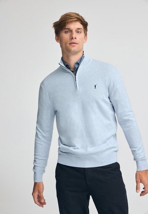 Lichtblauwe trui met een ritskraag, geribbelde manchetten en zoom, met een klein logo op de linkerkant van de borst, gedragen over een overhemd met kraag.