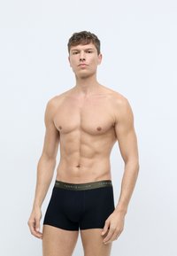 Sorte boxer shorts med en grøn talje, der har teksten "TOMMY HILFIGER". Modellen har kort hår og står foran en lys baggrund.