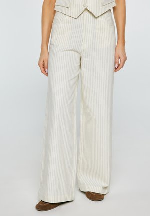 Kobieta nosząca beżowe spodnie w prążki o szerokich nogawkach w połączeniu z brązowymi butami, stojąca na tle jednolitego jasnego tła.