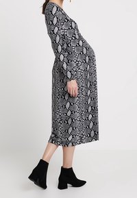 Kobieta w ciąży ubrana w midi sukienkę w czarno-szary wzór węża i czarne botki za kostkę, stojąca w profilu na jednolitym tle.