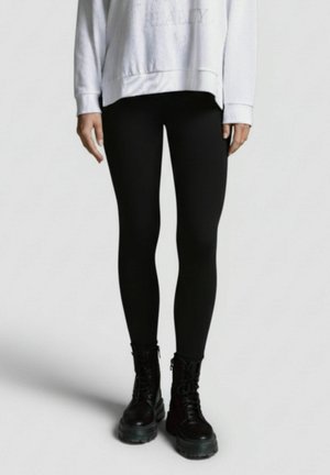 Schwarze Leggings aus dehnbarem Material, kombiniert mit klobigen schwarzen Stiefeln im Kampfstil. Das Oberteil ist ein locker sitzender weißer Sweatshirt.