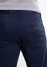 Námořnické modré bavlněné kalhoty s texturovaným povrchem, vybavené dvěma zadními kapsami, hnědým knoflíkovým detailem a poutky na pásek.