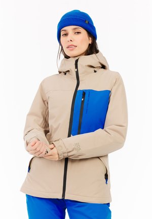 Beige waterproof jacket with blue chest panel, front zipper, adjustable cuffs, and hood, featuring pockets and logos. Model is wearing blue pants.