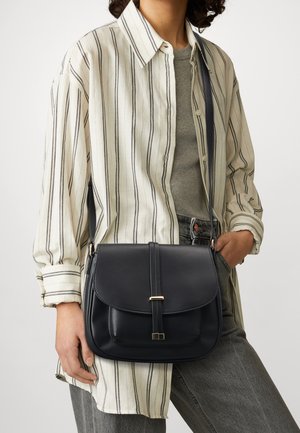 Femme portant une chemise rayée surdimensionnée et un pantalon gris, portant un sac en cuir noir en bandoulière avec un détail de fermoir doré.