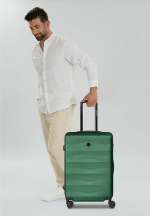 Hombre con camisa blanca y pantalones beige tirando de una maleta verde rígida con cuatro ruedas y un asa telescópica negra.