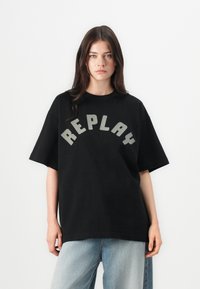 Svart oversized t-skjorte med korte ermer har stor grå "REPLAY" tekst i en buet design på forsiden. Glatt bomullsstoff.