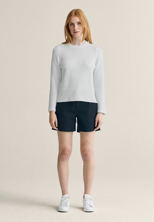 Ung kvinde med rødt hår står iført en tekstureret hvid langærmet trøje, sorte shorts og hvide sneakers mod en ensfarvet baggrund.