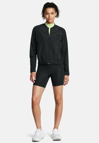 Schwarze Windbreaker-Jacke mit Reißverschluss vorne, strukturiertem Muster, elastischen Bündchen und Saum; kombiniert mit schwarzen Radlerhosen und weißen Sneakers.