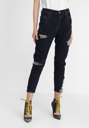 Kobieta nosząca czarne zniszczone dżinsy z postrzępionymi łatami oraz czarne botki na kostkę z ostrym noskiem i jasnżółtymi sznurowadłami.